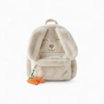 cartable en peluche beige en forme de tête de lapin, avec longues oreilles et broderie de visage souriant, décoré de deux carottes crochetées.