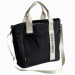 Cartable fourre-tout pour femme avec bandoulière de couleur noire avec la bandoulière, les anses et une bande blanches. Sur fond gris clair.