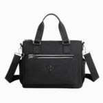 Cartable sac à main en nylon imperméable de couleur noire avec une bandoulière amovible, deux poches à l'avant et un grand espace principal. Sur fond gris clair.